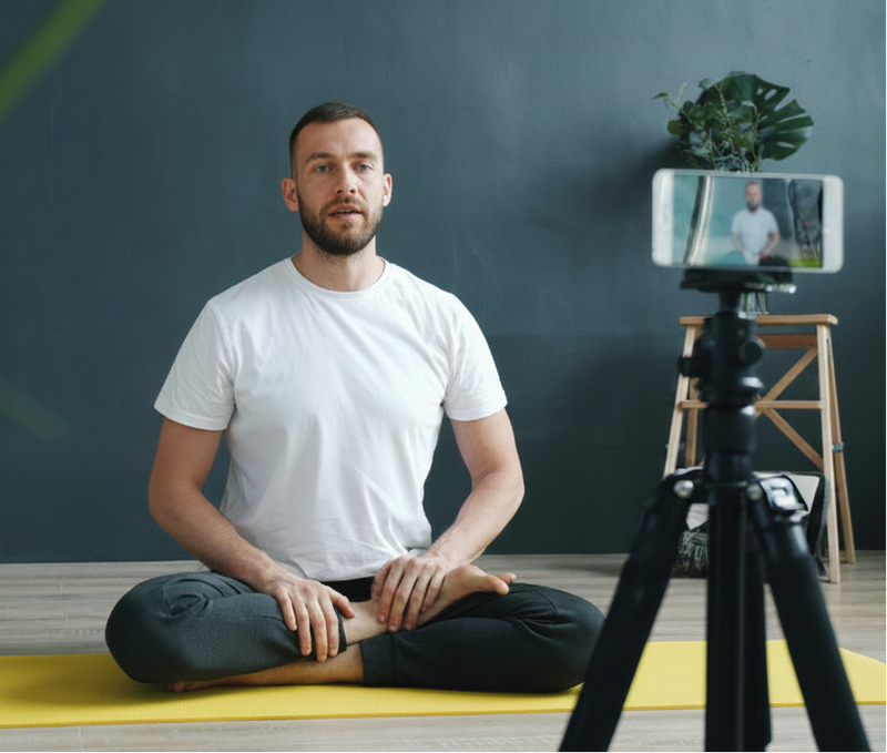 Man sitting on a yoga mat with a camera on a tripod recording him.