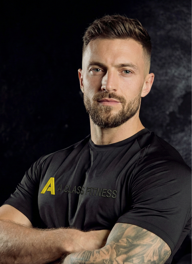 Man wearing a black 'A Class Fitness' t-shirt with tattoos on arms against a dark background