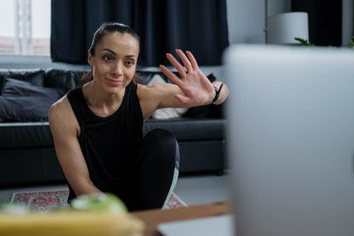 Woman in black athletic wear sitting on a yoga mat in a living room, waving at a laptop.