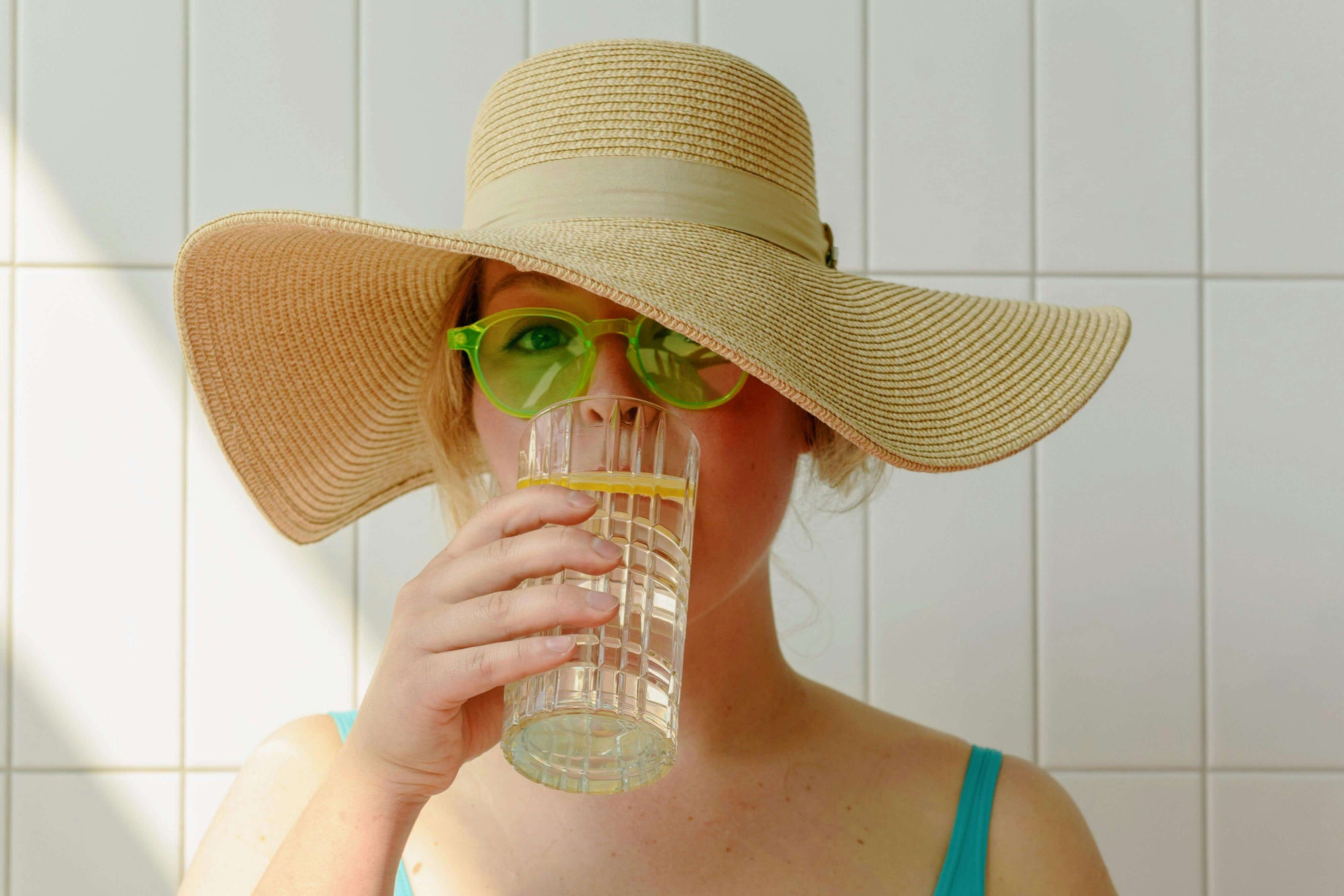 Person wearing a wide-brimmed hat and sunglasses, holding a glass against a tiled wall.